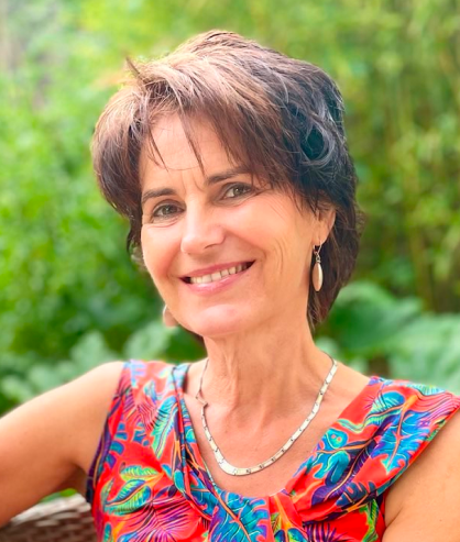 Heike lächelt in die Kamera. Sie sitzt. Im Hintergrund sieht man viele grüne Pflanzen. Sie trägt eine schlichte Kette und Ohrringe. Sie hat ein rotes Kleid mit abstrakten, bunten Blättern an.